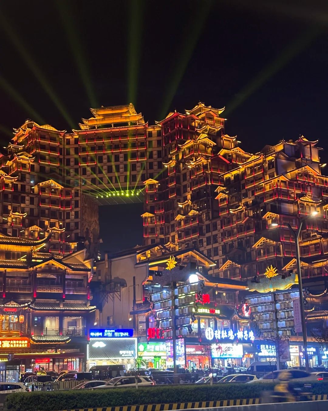 72 Qilou lit up at night in Zhangjiajie, photographed from the opposite side of the avenue, with a crash barrier, moving traffic and shopfronts in the foreground.