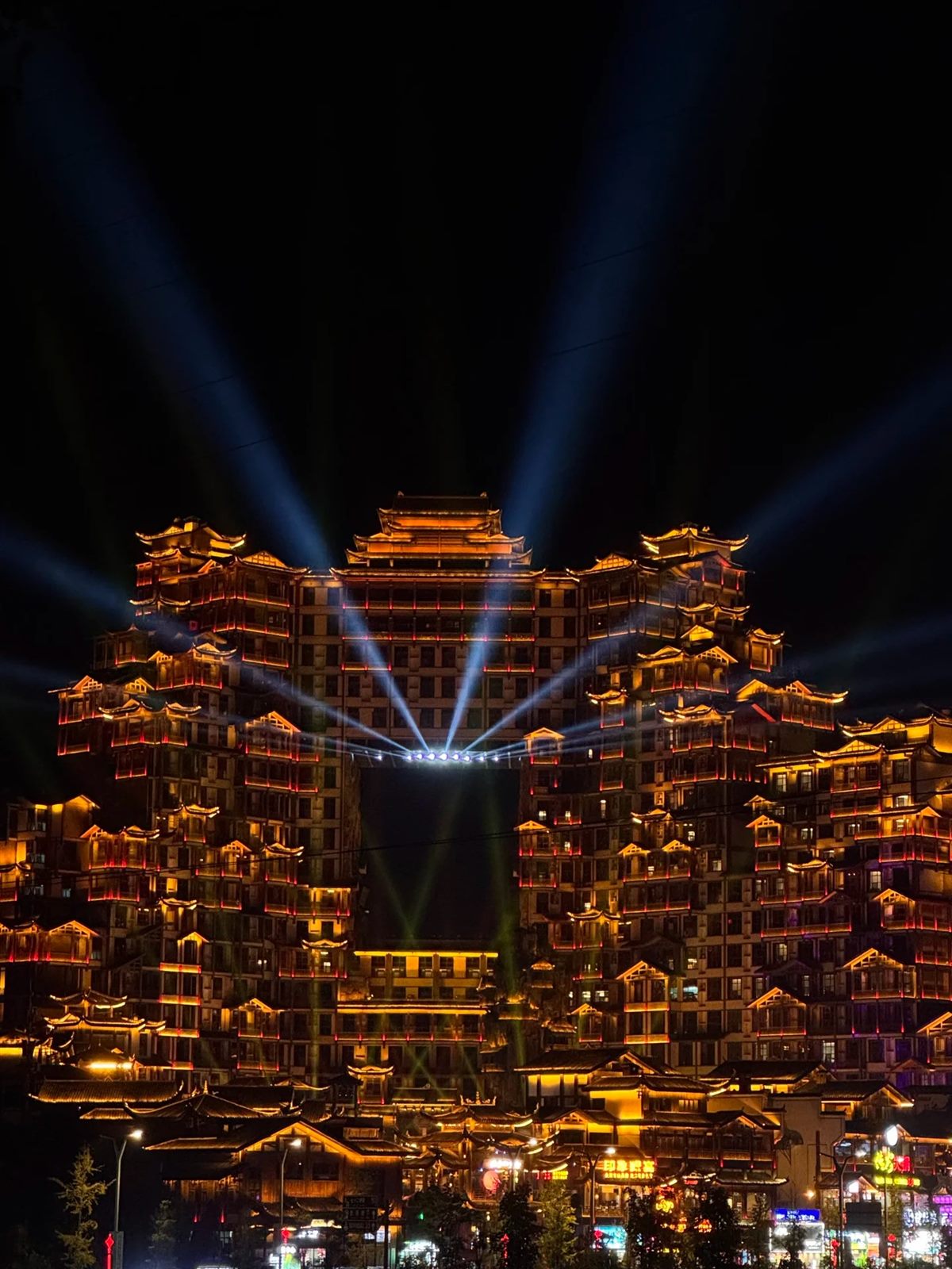 The full 72 Qilou complex in Zhangjiajie lit at night, tiers of timber balconies picked out in orange and red with searchlight beams above, and lit shopfronts along the street below.