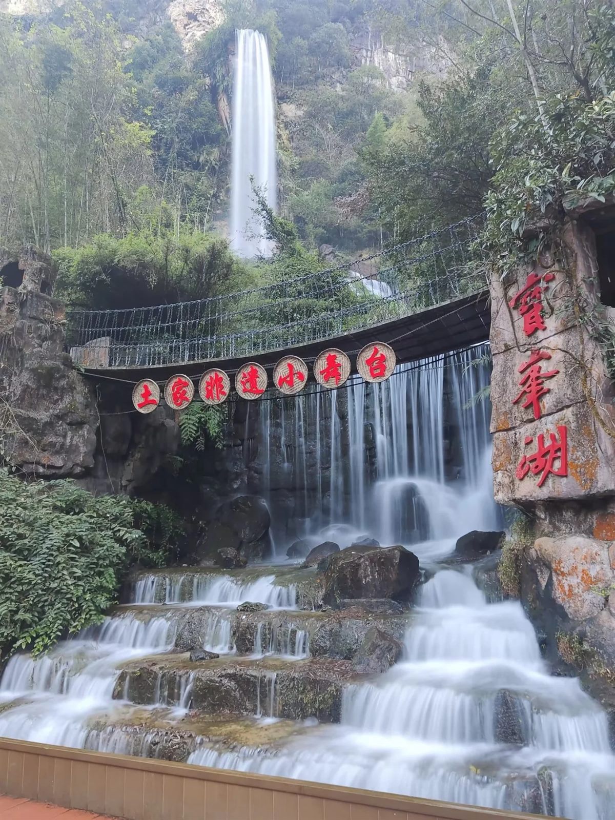 The waterfall and stepped cascade beside the Baofeng Lake name stele, with a footbridge crossing above it.