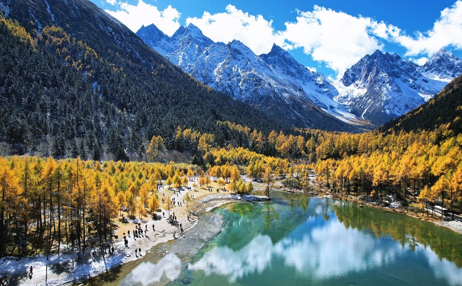 Aerial view of a still lake with cloud and snow-covered peaks mirrored across its surface, golden larch along both banks and a pale, crowded shoreline.