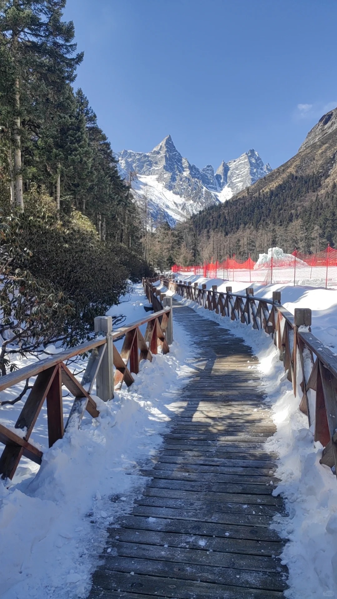 A timber boardwalk with post-and-rail fencing running away through snow towards jagged snow-covered spires at the head of the valley, orange safety netting and a snow mound to the right.