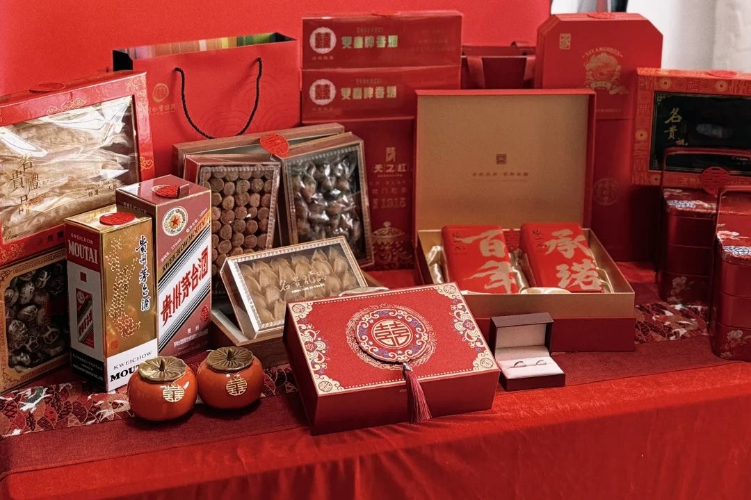 A table of red betrothal gifts laid out: spirits, cigarettes, cake boxes, dried-seafood hampers, tea caddies and an open ring box