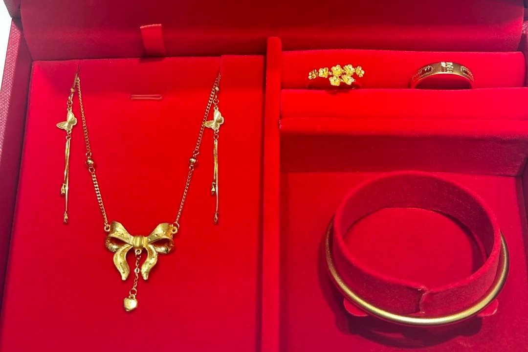 A red velvet presentation box holding a gold necklace with a bow pendant, two gold rings and a gold bangle