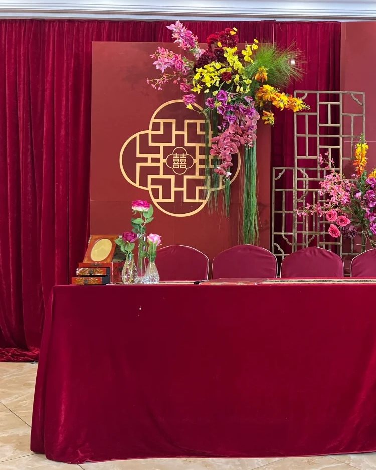 A Chinese wedding sign-in desk in deep red velvet, with a guest register, an ink-seal box and flowers, in front of a double-happiness board
