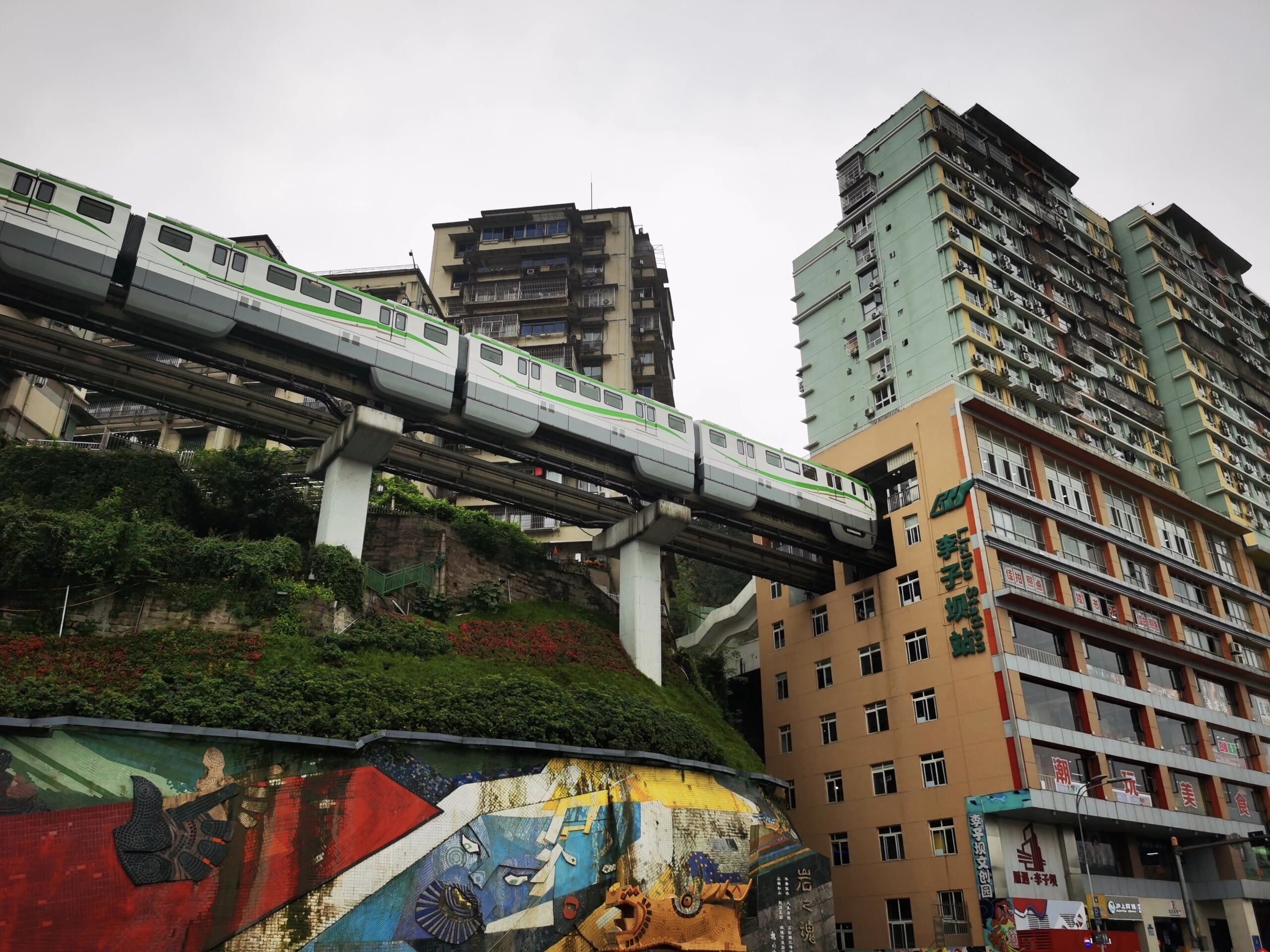 A Chongqing Metro Line 2 monorail train passing through the middle floors of a residential apartment block at Liziba station — full train mid-traversal, dense apartment blocks behind, street-art mural in the foreground.