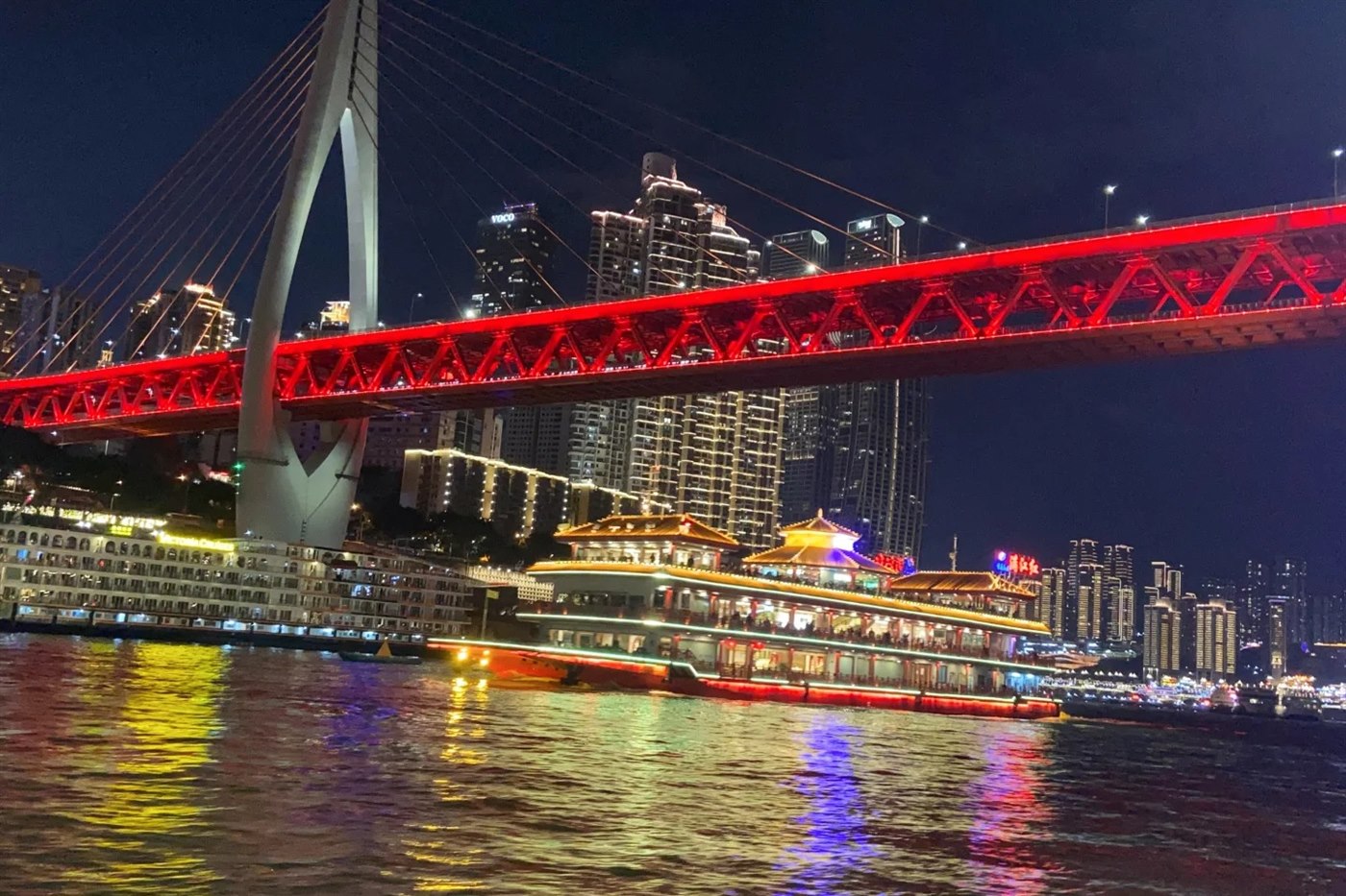 A double-deck sightseeing boat with pagoda-style roofs, lit and full of passengers, passing beneath the Qiansimen bridge at night; the bridge's red steel truss runs across the top of the frame and the city towers rise behind.