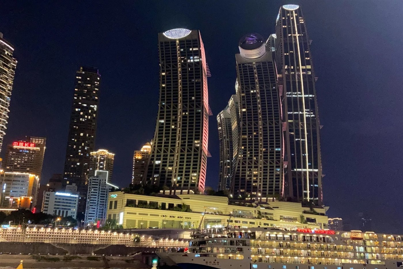 The Raffles City towers at Chaotianmen in Chongqing lit at night, with a large multi-deck river cruise ship lettered CENTURY LEGEND berthed at the terminal below them.
