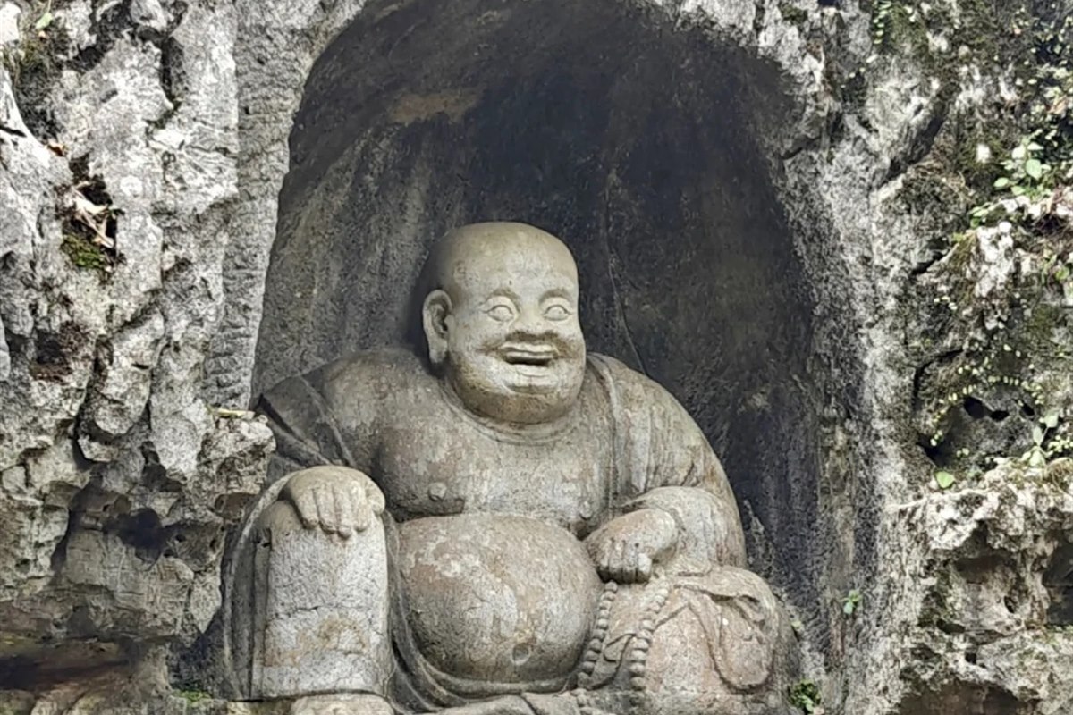 The large stone laughing Maitreya Buddha carved into a rock niche at Feilai Feng, Hangzhou, round-bellied and smiling, with prayer beads in one hand.