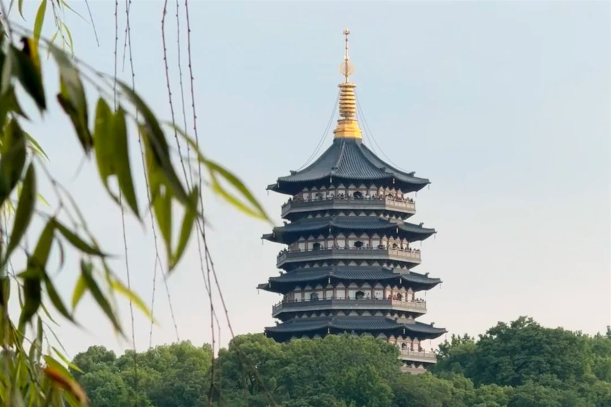 Leifeng Pagoda in Hangzhou by day, seen through willow branches — five tiers of wide dark eaves under a gilded finial, with visitors standing on the open galleries.
