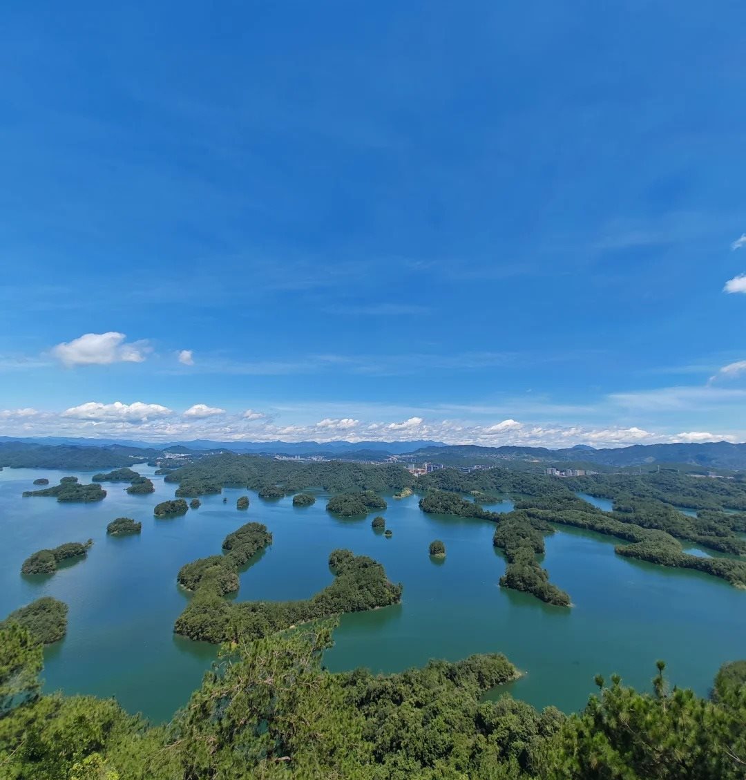 Wide view from the Huangshanjian summit at Qiandao Lake over dozens of green islands scattered in deep blue water, with a town visible on the far shore