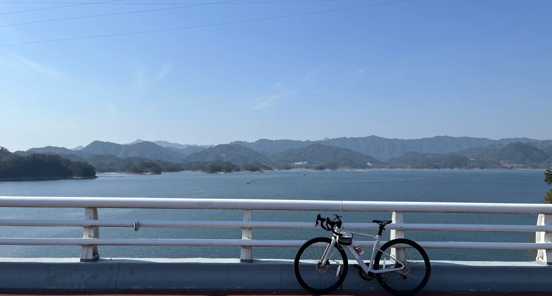 A bicycle propped against the white railing of a bridge on the ring-lake road at Qiandao Lake, with a marked red cycle lane and layered green hills across the water