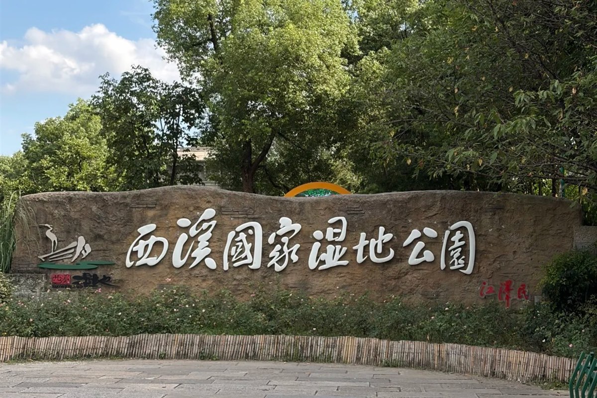 The rough stone entrance sign at Xixi National Wetland Park in Hangzhou, carved with the park's Chinese name in large white characters, backed by trees under a blue sky.