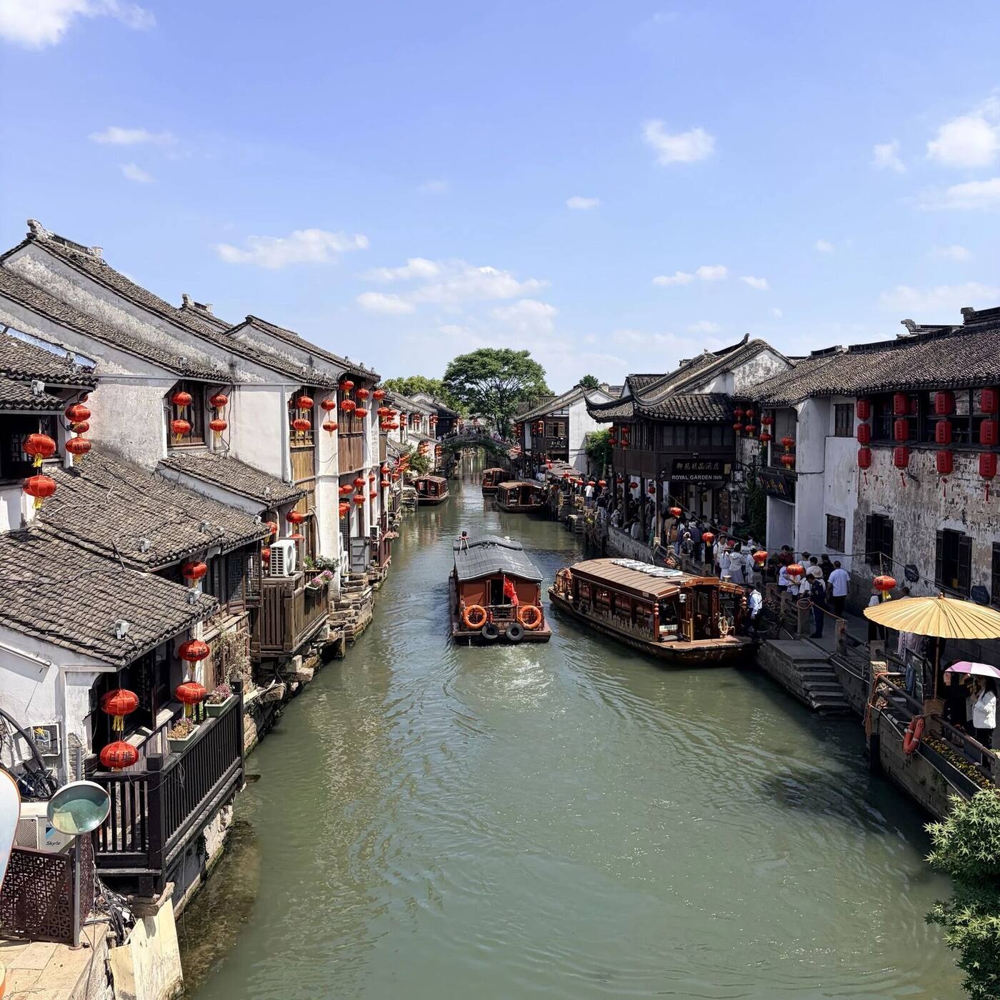 A Suzhou canal street lined with whitewashed houses and red lanterns, with wooden tour boats moored along the water.