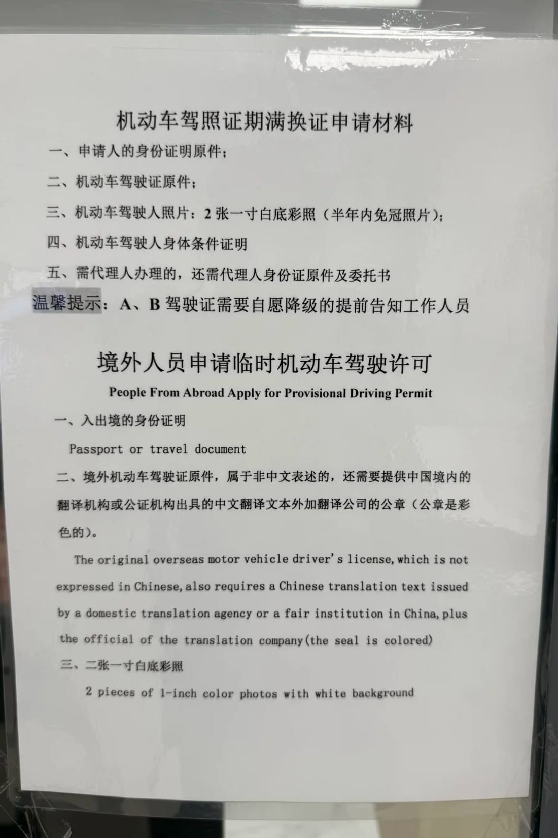 Bilingual notice posted at the Beijing Daxing vehicle administration desk headed 'People From Abroad Apply for Provisional Driving Permit', listing a passport or travel document, the original overseas licence with a Chinese translation bearing the translation company's colour seal, and two 1-inch white-background photos.