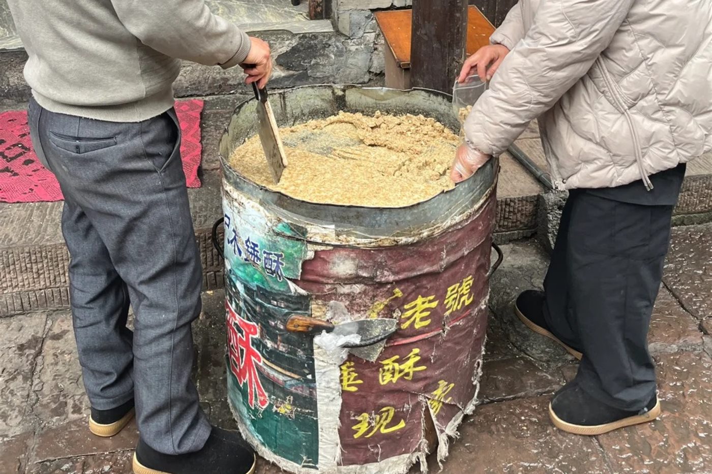 Two stallholders working a batch of ginger candy in a metal drum on a Fenghuang lane, one holding the blade used to break it up, the drum painted with the words for mallet-pounded ginger crumble.