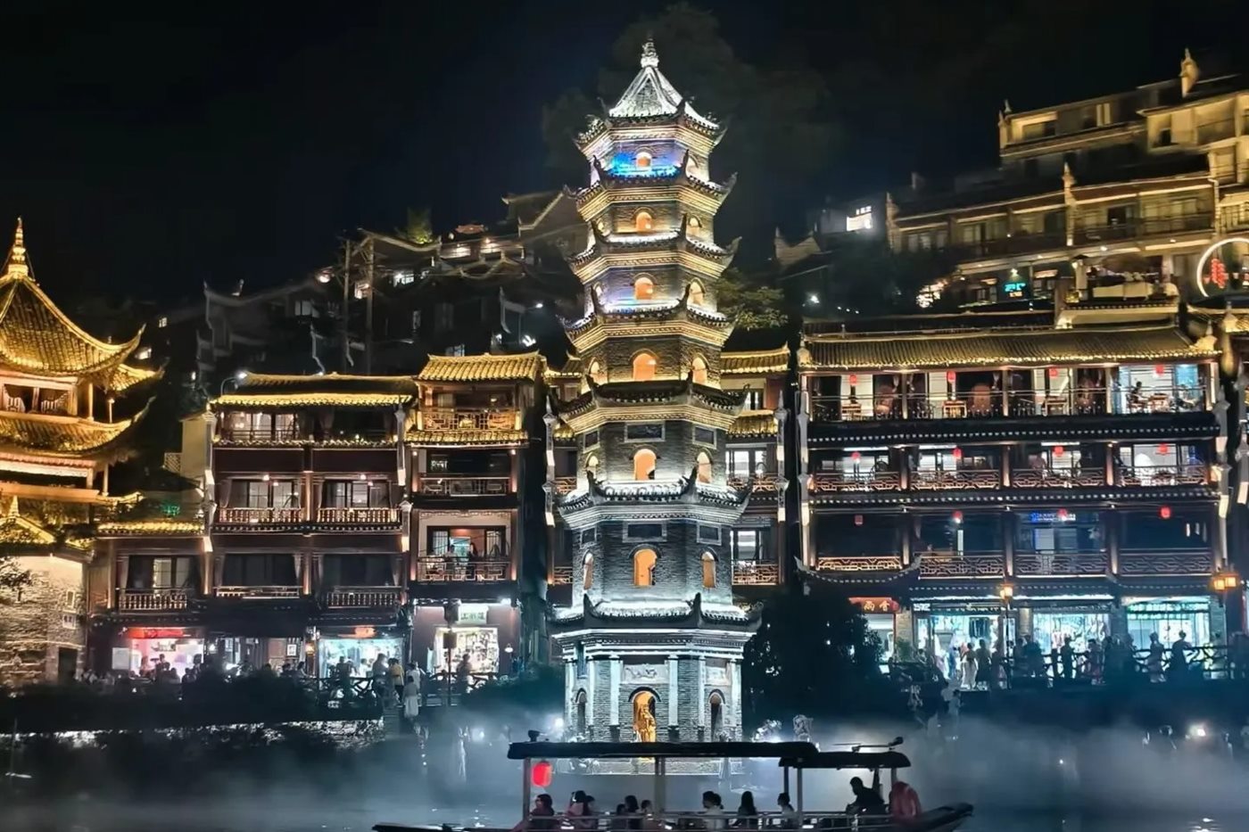 A lit passenger boat on the Tuojiang River in Fenghuang at night, passing below the illuminated Wanming Pagoda with the riverfront buildings glowing behind.