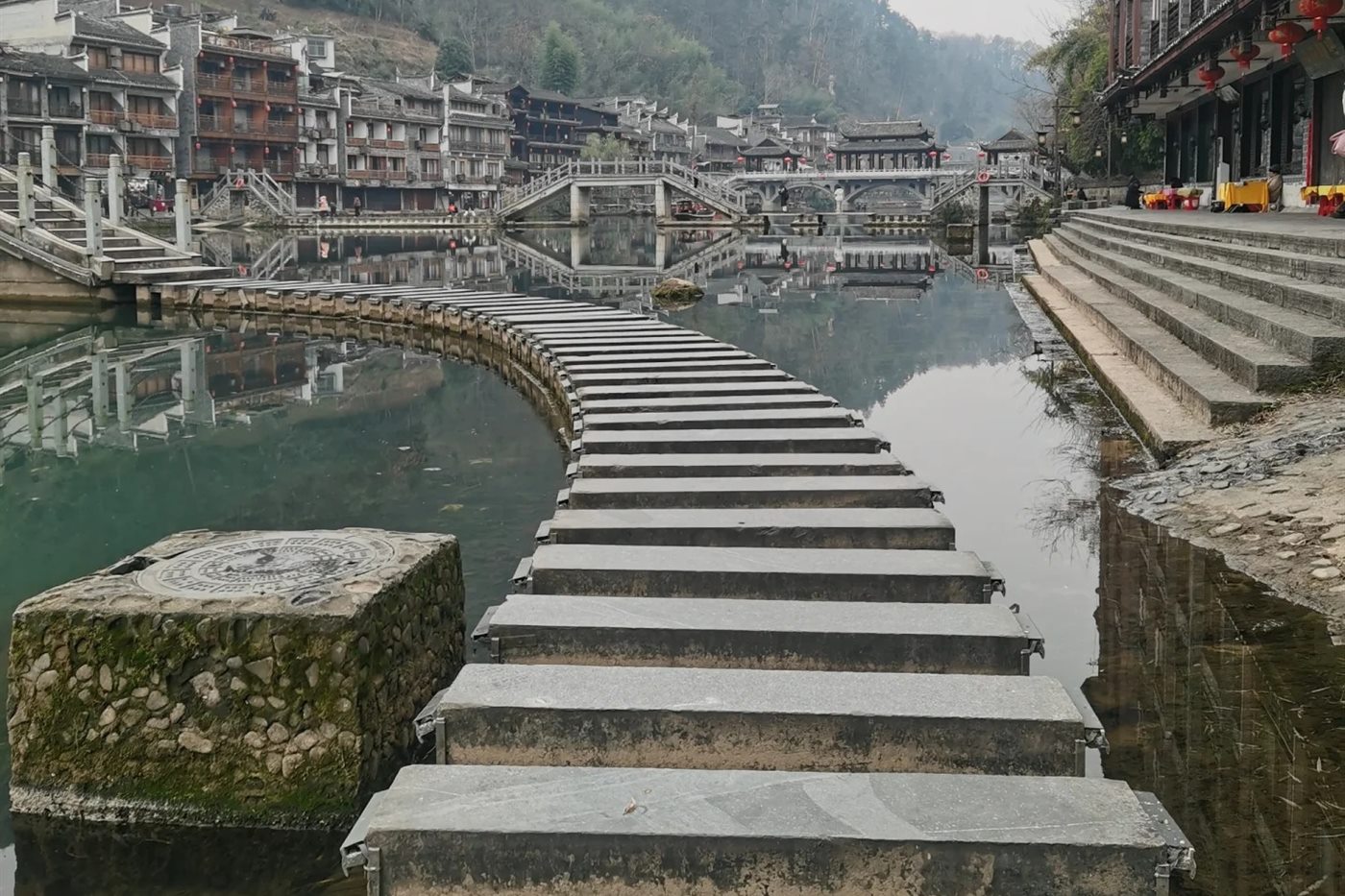 The stepping stones of Fenghuang crossing the Tuojiang River empty of people, with the arched bridges and stilt houses of the old town behind.