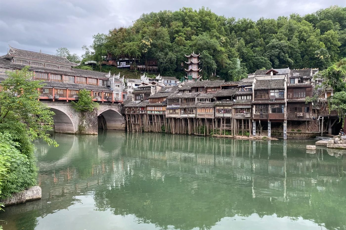 Wooden stilt houses of Fenghuang Ancient Town standing on timber piles over the green Tuojiang River, with an arched covered bridge on the left and a pagoda on the wooded hill behind.