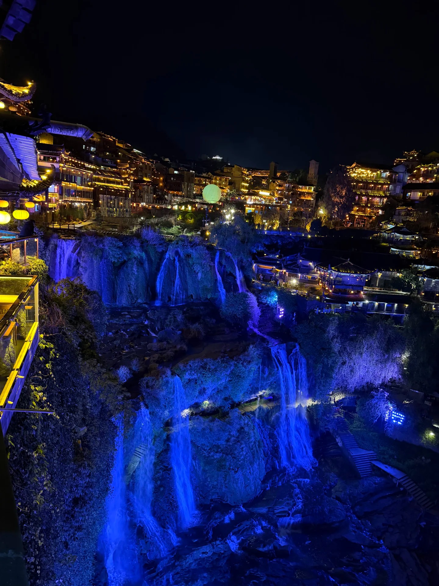 Furong Ancient Town at night — the waterfall lit blue below the warm-lit stilt houses and red lanterns of the old town.
