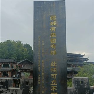 The Xizhou Bronze Pillar stele in Furong Ancient Town, dark metal with gold-inscribed Chinese characters, Tujia stilt houses behind.