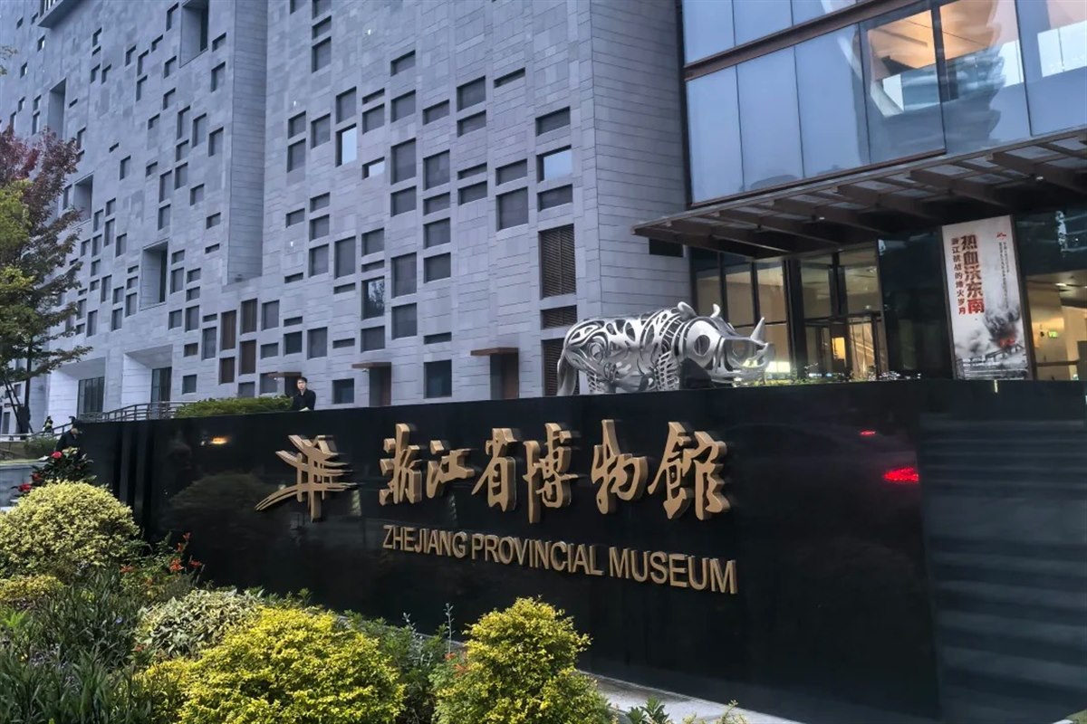 The bilingual entrance sign of the Zhejiang Provincial Museum's Zhijiang building, with the museum's stone-and-glass facade behind it.