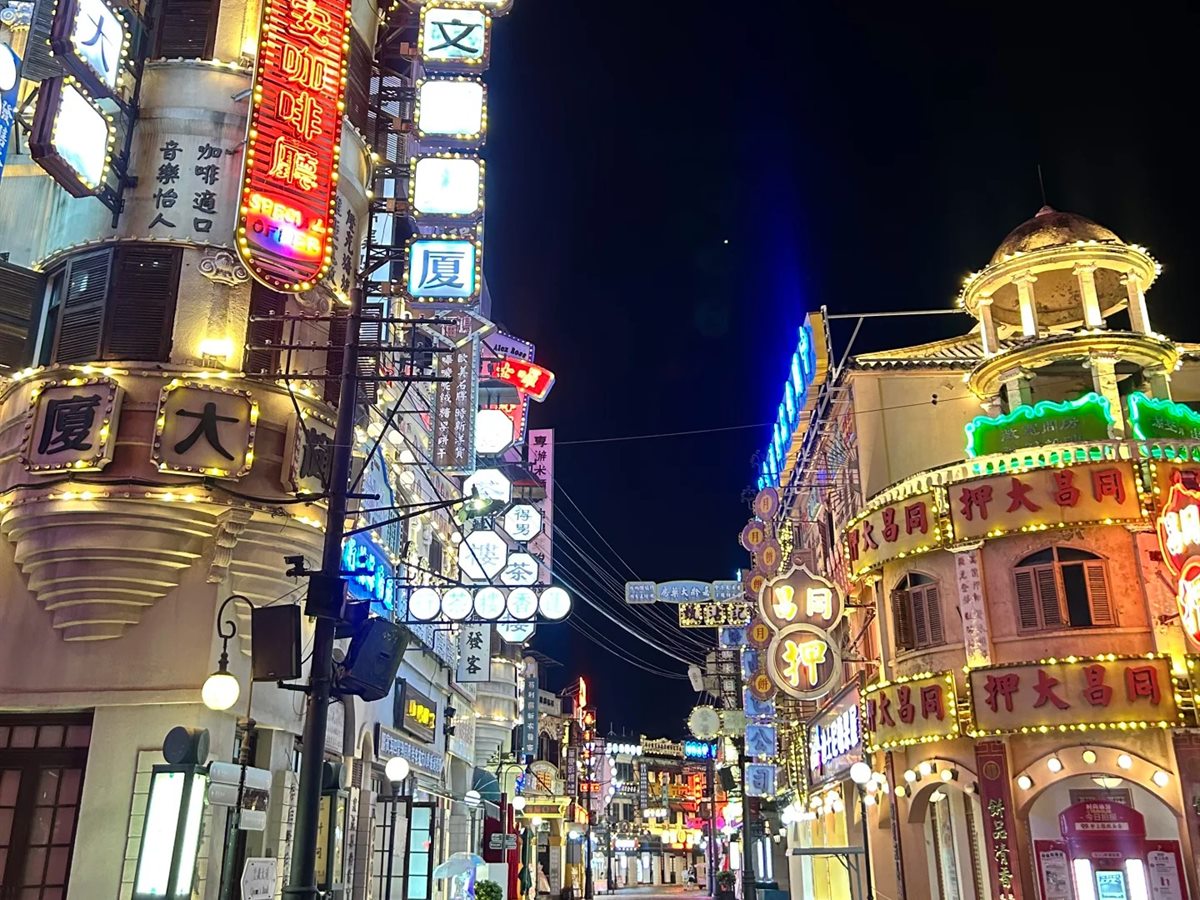 A cobbled street on Hengdian's Guangzhou Street · Hong Kong Street set at night, lined with Republican-era shopfronts whose facades are stacked three storeys high with lit neon and bulb-lit signs in Chinese, including a pawnbroker's and a coffee house, receding into the dark.