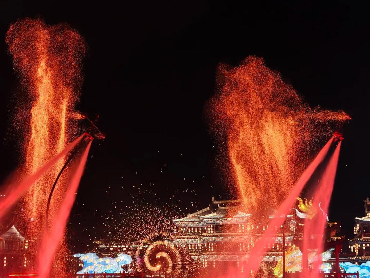 The molten-iron show at Hengdian after dark: two smiths silhouetted against the night fling arcs of liquid iron that burst into towering orange showers of sparks above a stretch of water, with lit Song-dynasty halls and a glowing spiral of fire behind them.