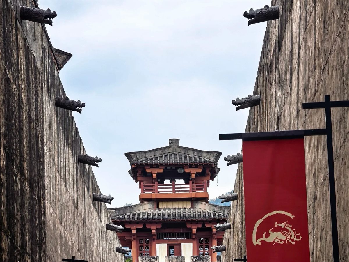A narrow canyon between two enormous grey rammed-earth and stone set walls at Hengdian World Studios, carved dragon-head spouts projecting from both parapets, red banners bearing a gold dragon hanging from posts, and a red-and-grey timber gate tower closing the far end under an overcast sky.