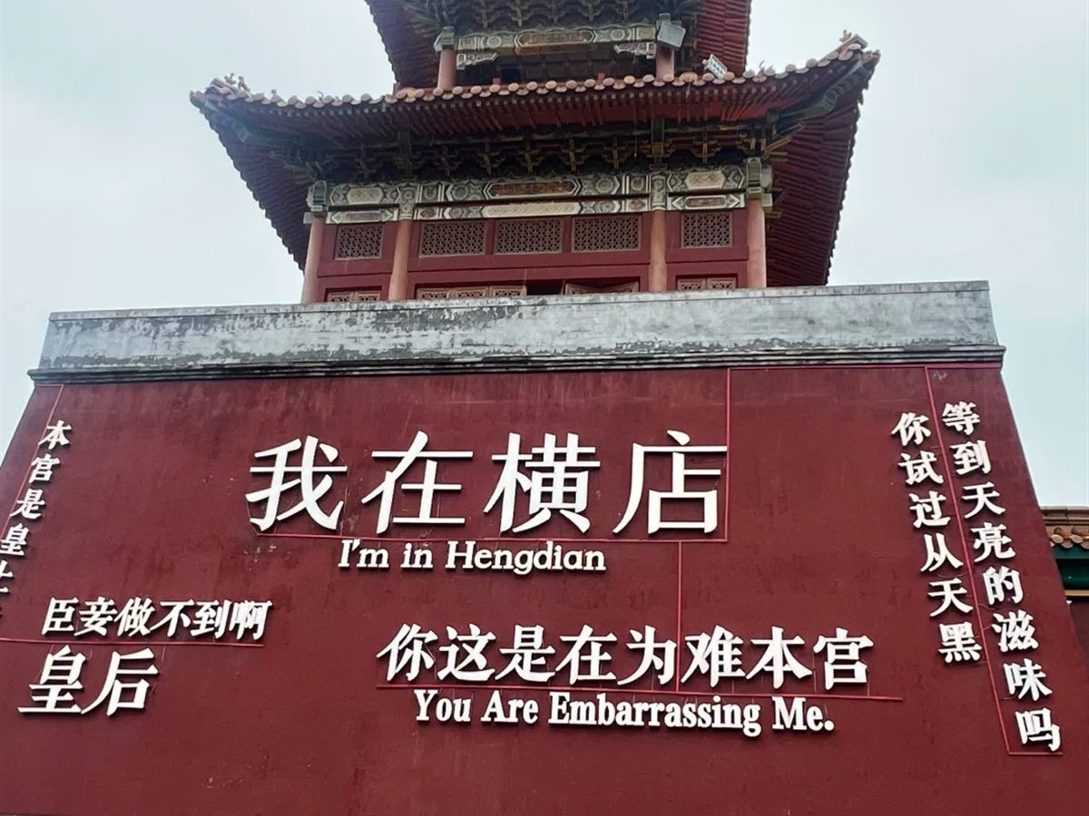 A large dark red wall at Hengdian World Studios carrying the raised characters 我在横店 with 'I'm in Hengdian' beneath them, and smaller palace-drama catchphrases in Chinese and English including 'You Are Embarrassing Me.', with a two-tiered red and gold pavilion rising behind the wall.