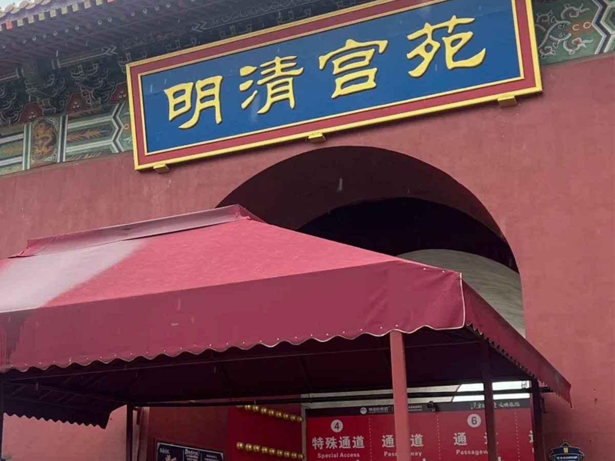 The entrance to Ming and Qing Palatial Garden at Hengdian: a blue and gold name board reading 明清宫苑 mounted on a dark red gatehouse above an arched opening, with a red canopy below sheltering numbered entry lanes whose signs read 特殊通道 Special Access and 通道 Passageway.