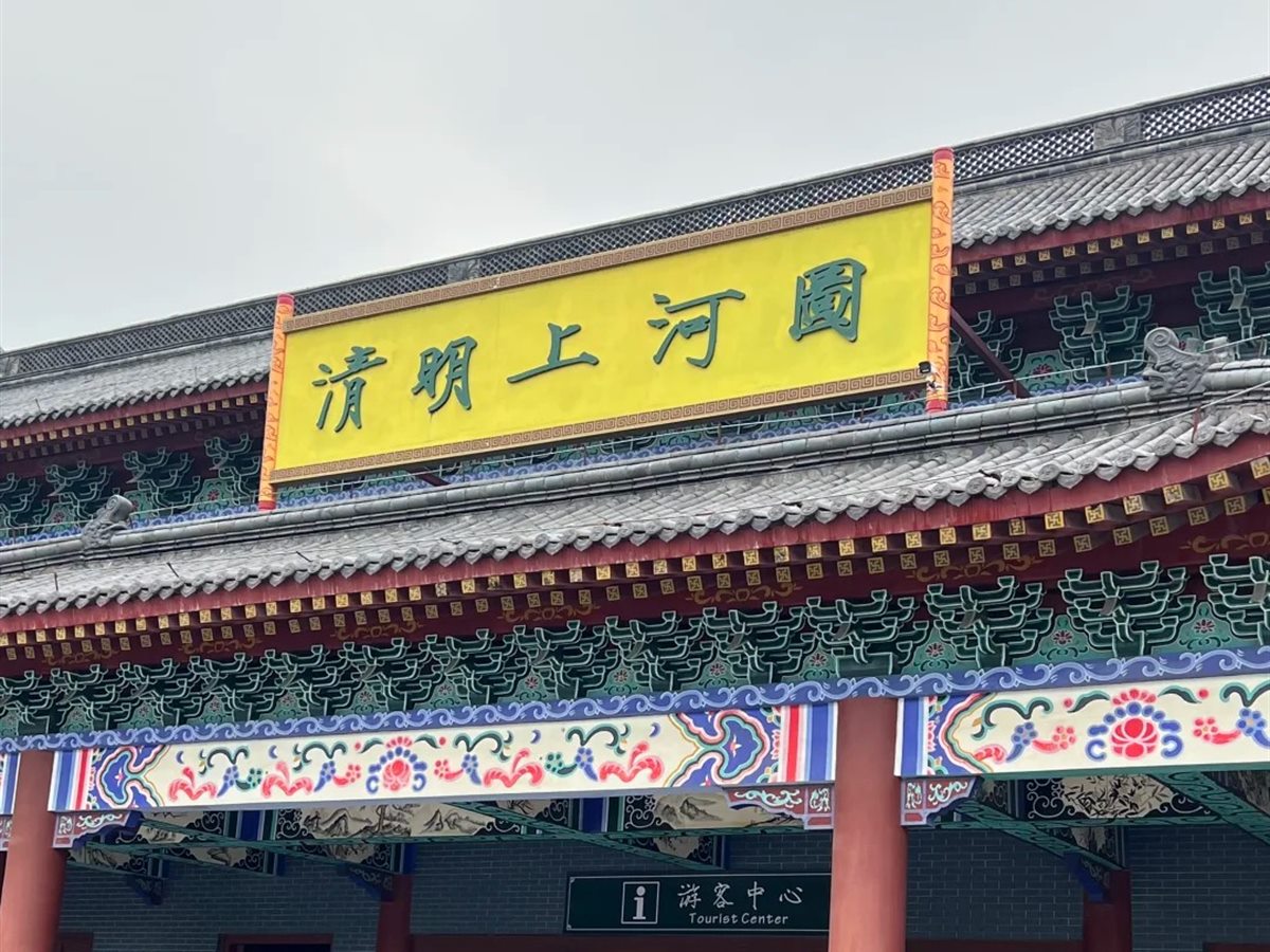 The entrance to Qingming Shanghe Tu at Hengdian: a grey-tiled gatehouse with red columns and green-and-blue painted beams, carrying a yellow name board lettered 清明上河圖 in green, with a Tourist Center sign and a rack of hired costumes visible in the shade beneath.
