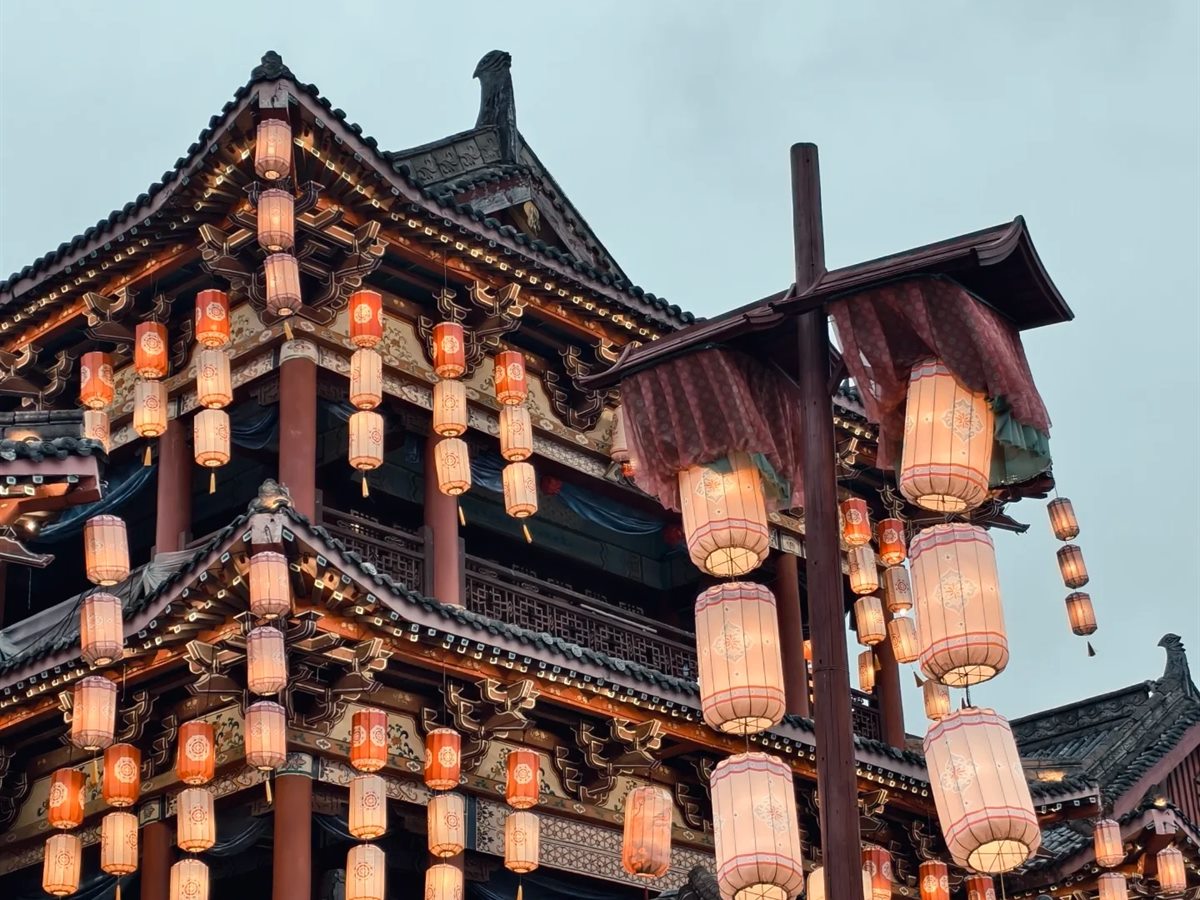A multi-storey Song-dynasty timber building at Qingming Shanghe Tu photographed at dusk against a pale grey sky, its balconies and eaves hung with several hundred lit orange and white lanterns, with a tall lamp post carrying a further cluster of lanterns in the foreground.