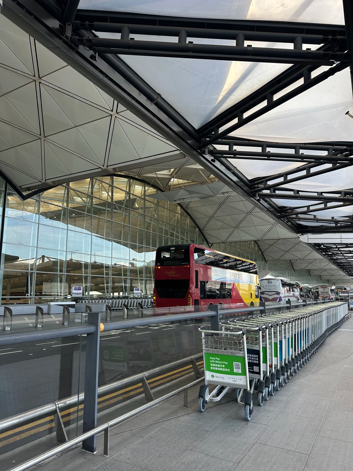Terminal 1 curbside at Hong Kong International Airport under the white lattice canopy, with a red CityFlyer double-decker airport bus and rows of luggage trolleys