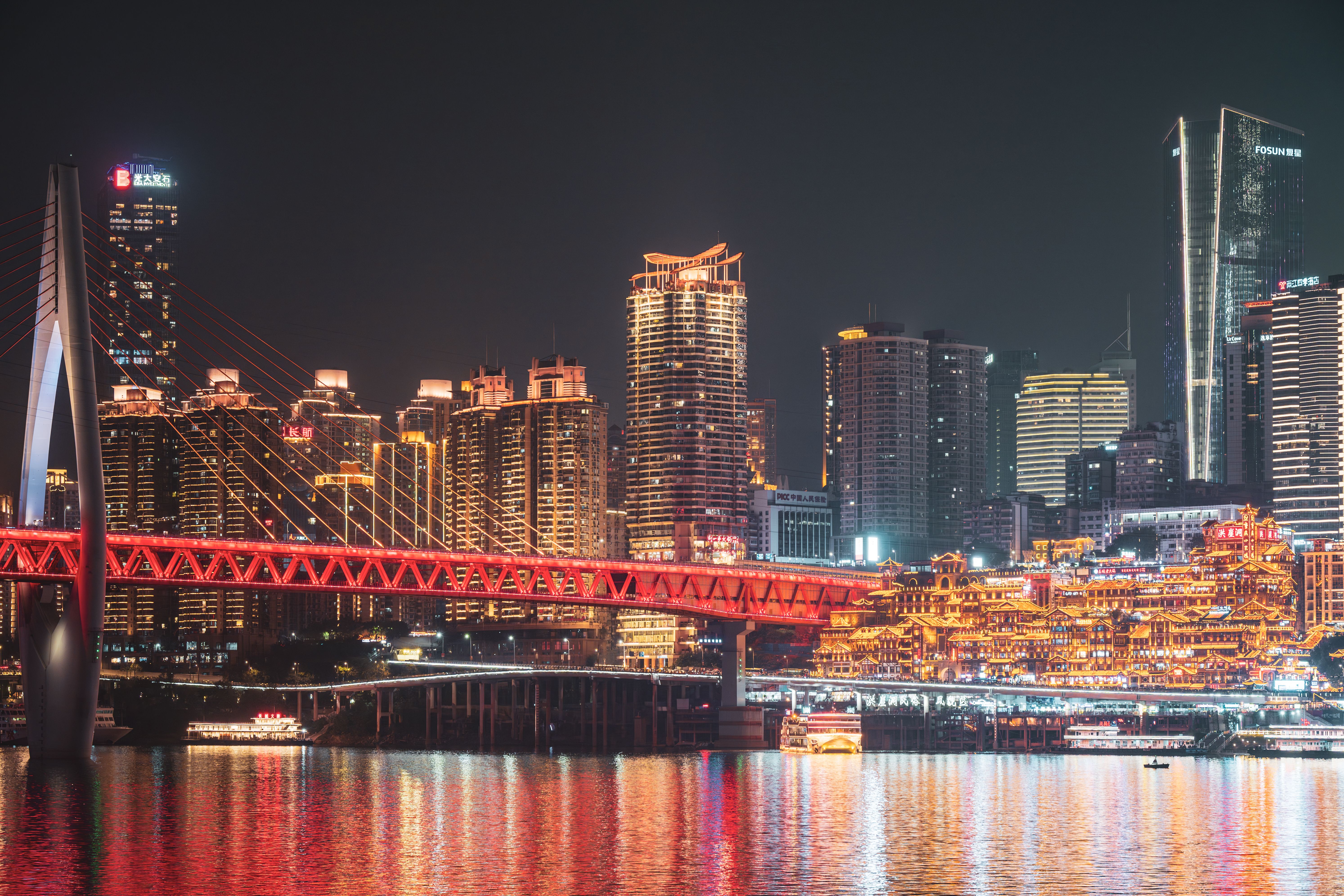 Hongyadong lit up at night seen from across the Jialing River, with the red Qiansimen Bridge spanning the foreground, Chongqing.