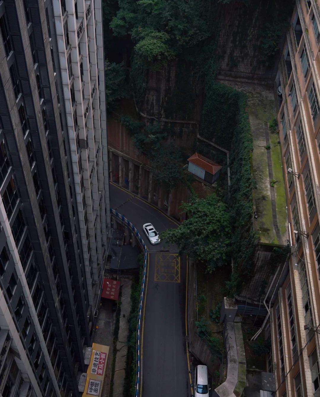 Looking straight down from height into a narrow Chongqing street canyon between two residential towers, with a hairpin road and a single white car far below.