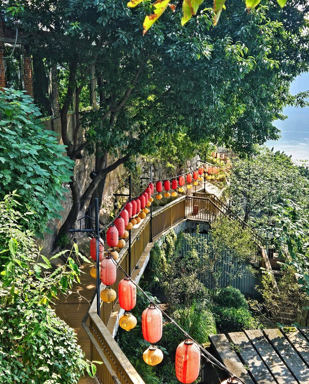 The Shancheng Lane cliff boardwalk seen from above, curving downhill under a long string of red and yellow lanterns, with a stone retaining wall on one side and the hazy river below on the other.