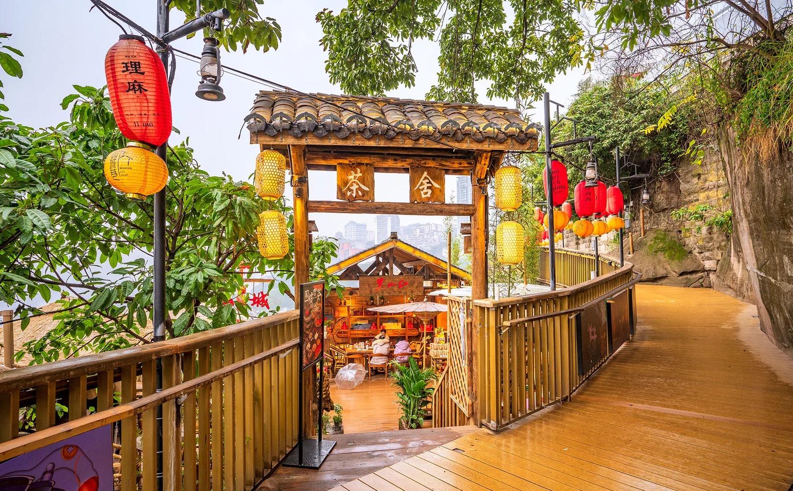 A timber tea-house gateway signed 茶舍 opening off the Shancheng Lane boardwalk, hung with red and yellow lanterns, one of which reads 山城巷 / Mountain City Alley; the city towers show hazily beyond the cliff edge.