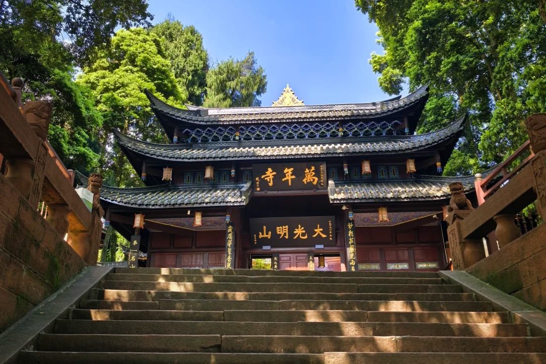 The entrance gate of Wannian Temple on Mount Emei, seen from the foot of its stone stair, with two gold-lettered plaques on the dark timber front.