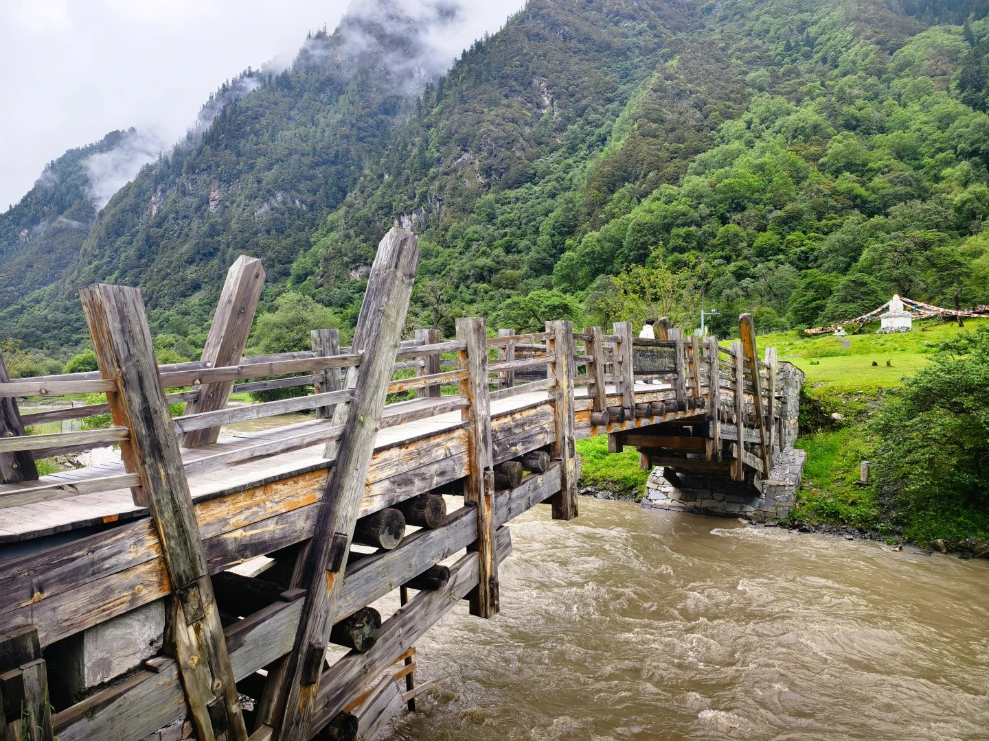 A timber bridge over a fast grey-brown river in Shuangqiao Valley, forested slopes rising into low cloud on both sides, and a white stupa hung with prayer flags on the far bank.