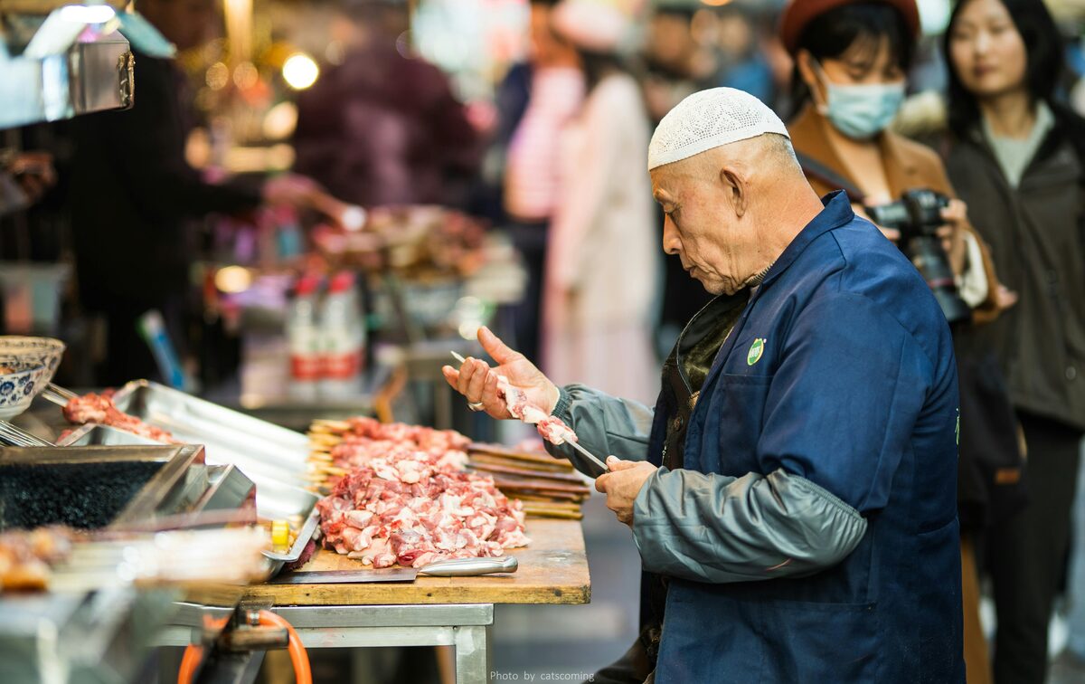 A Hui Muslim vendor in a white skullcap preparing lamb skewers at a stall in Xi'an's Muslim Quarter.