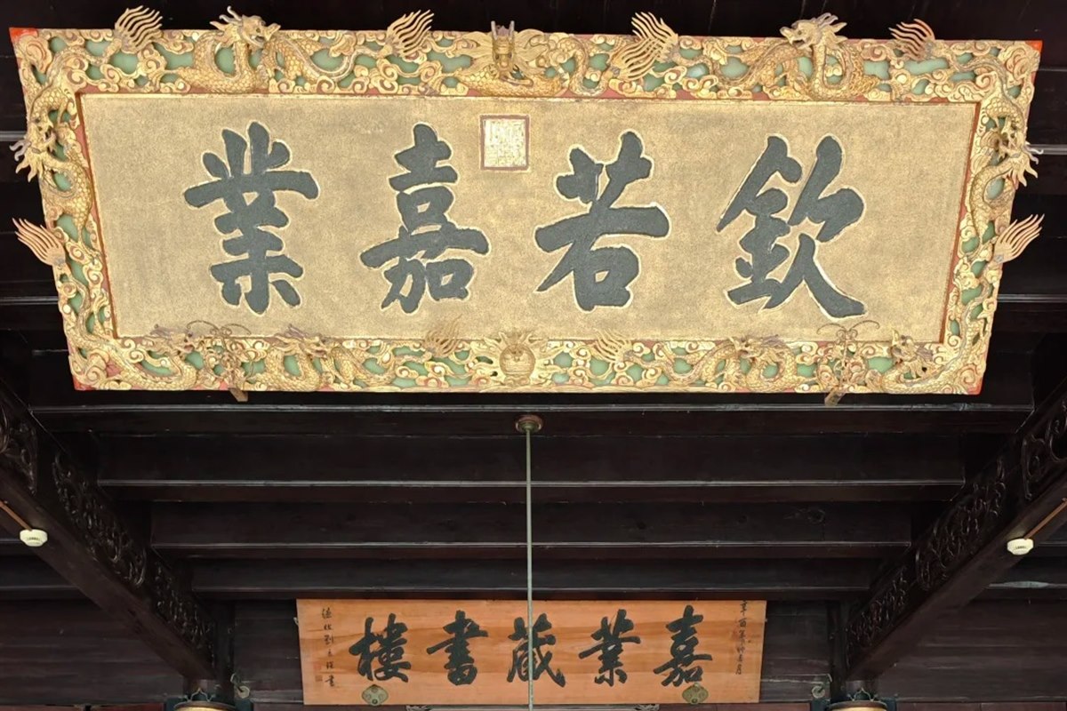 The main hall of the Jiaye Library in Nanxun: a gilded imperial plaque reading Qin Ruo Jia Ye in a dragon-carved frame hangs above a smaller board reading Jiaye Cangshulou, over dark rosewood chairs and a round table.