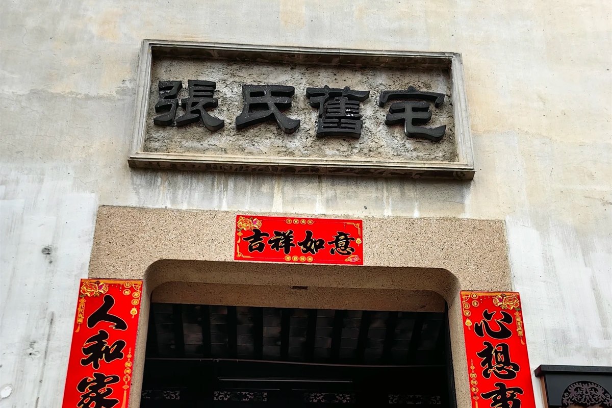 The entrance to the Zhang family's former residence in Nanxun: a plain rendered wall with a carved stone-framed plaque reading Zhang Shi Jiu Zhai, red New Year couplets down each side of the doorway, and a lantern-hung courtyard beyond.