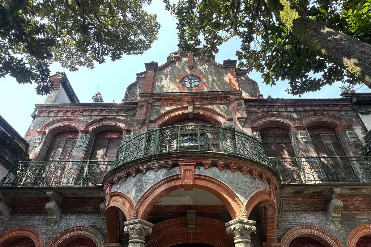 The European wing of Zhang Shiming's house in Nanxun: a red-and-grey brick baroque façade with a curved wrought-iron balcony, a rose window in the gable and Corinthian columns below, seen from the courtyard through overhanging branches.