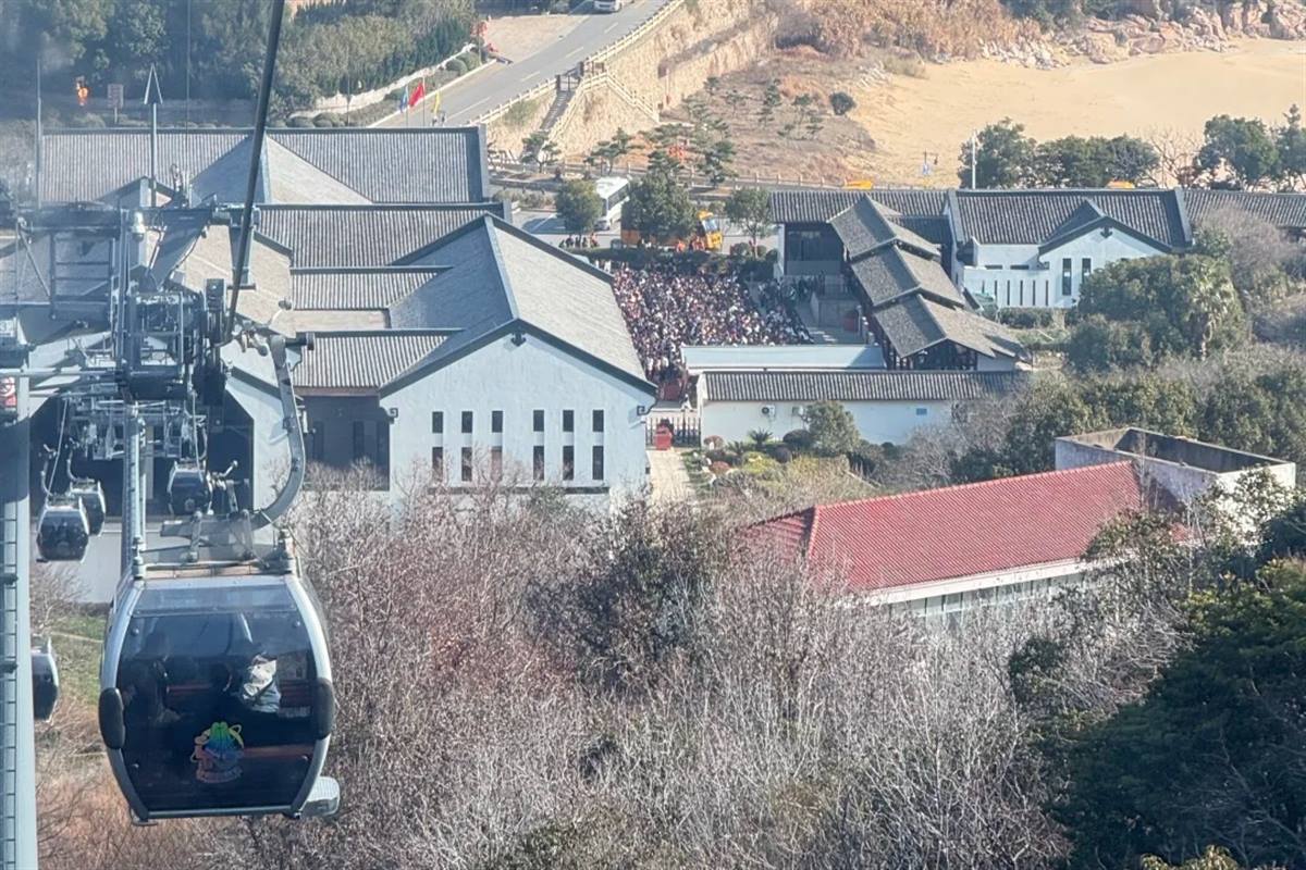 A gondola of the Foding Shan cable car descending over bare winter trees on Putuoshan, with temple halls, a road and a dense queue of visitors at a temple gate on the slope below.