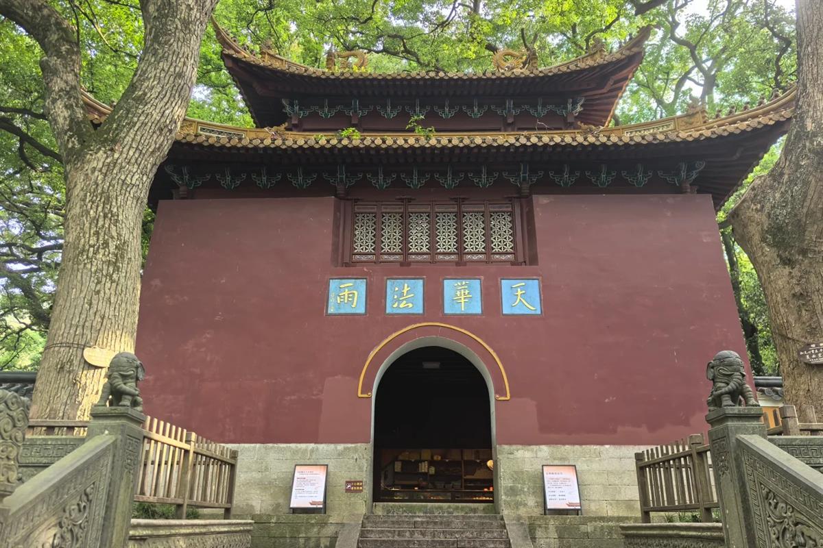 The Tianwang hall of Fayu Temple on Putuoshan: a dark red wall beneath a double-eaved tiled roof, four gold characters on blue panels above an arched doorway, framed by two old camphor trees.