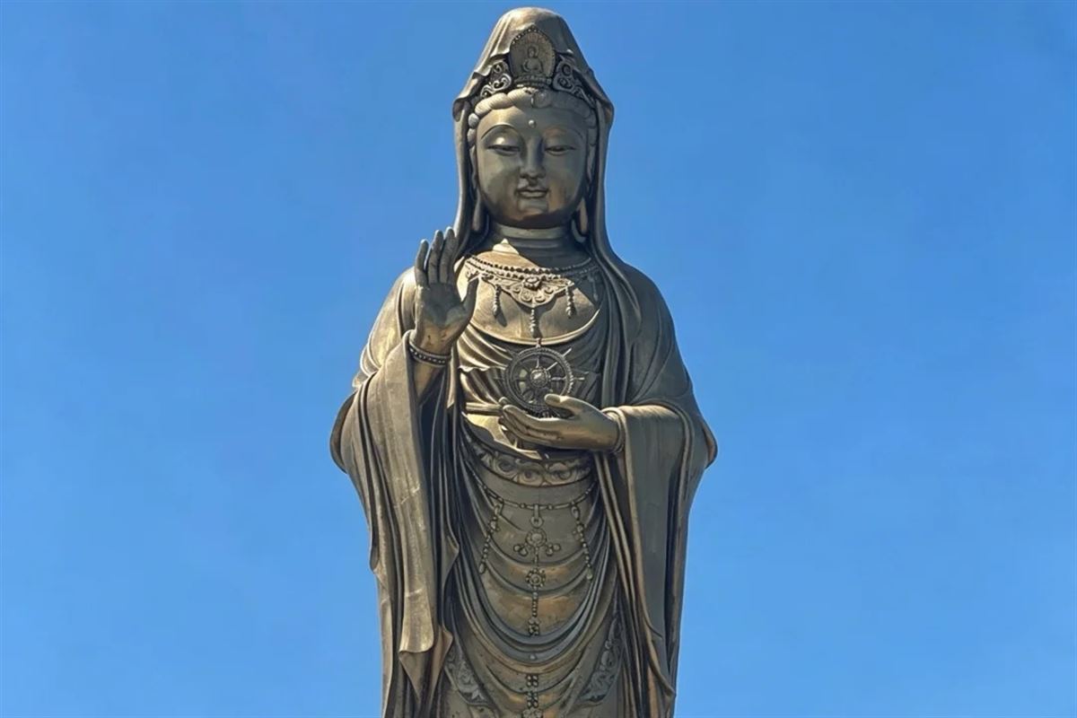 The bronze Nanhai Guanyin statue on Putuoshan against a clear blue sky: a standing figure in flowing robes with the right hand raised and a dharma wheel held at the chest.