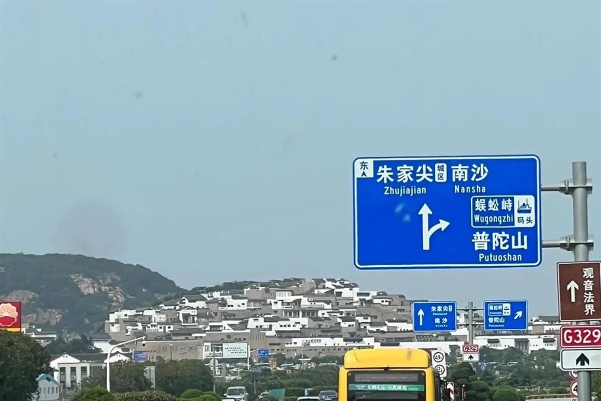 A blue Chinese motorway direction sign on Zhujiajian: Zhujiajian and Nansha straight ahead, and Wugongzhi pier with Putuoshan signposted to the right, above traffic and a hillside covered in white buildings.