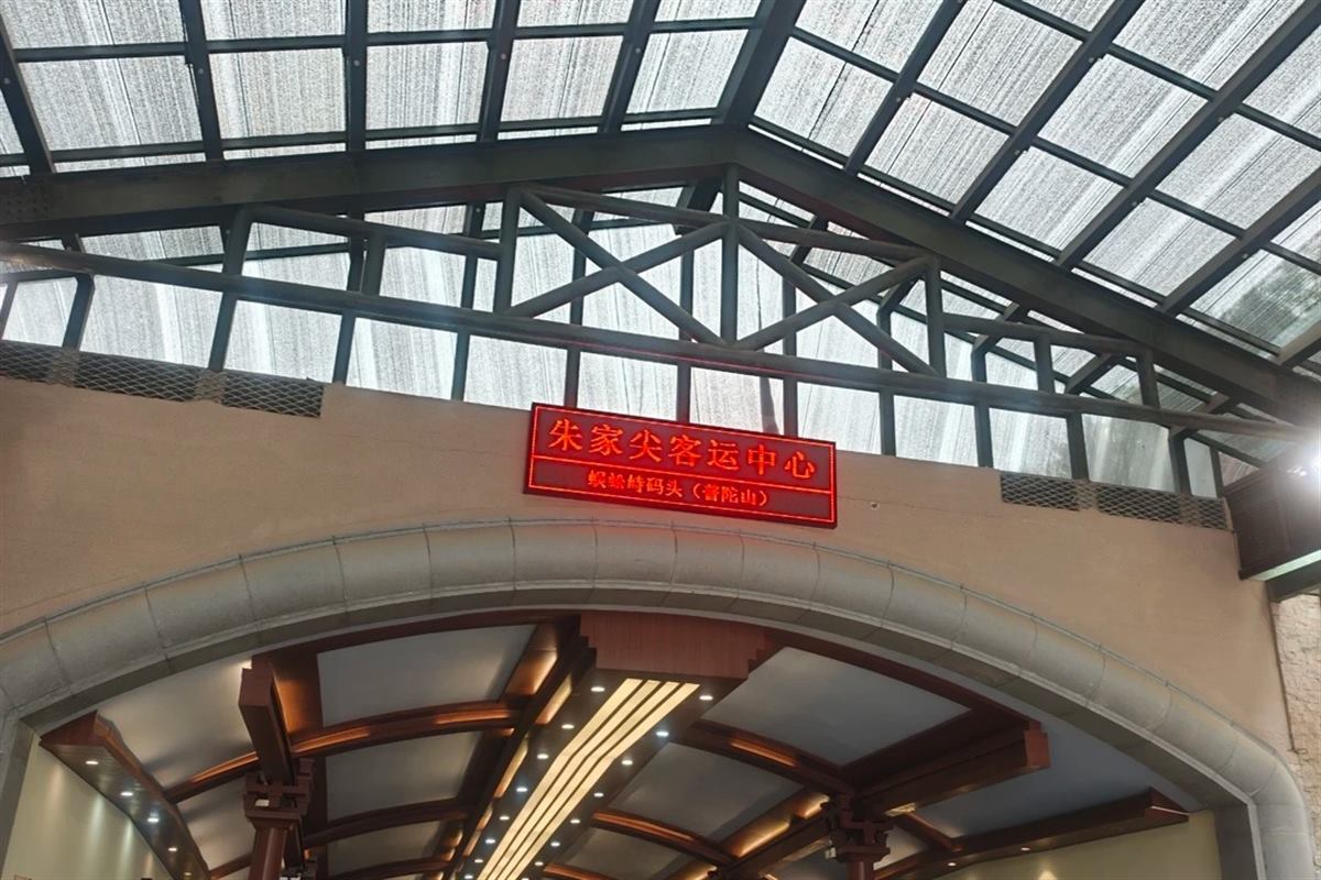 The glass-roofed concourse of the Zhujiajian passenger transport centre, with a red LED sign reading 朱家尖客运中心 and beneath it 蜈蚣峙码头(普陀山)above the arched entrance to the boarding corridor.