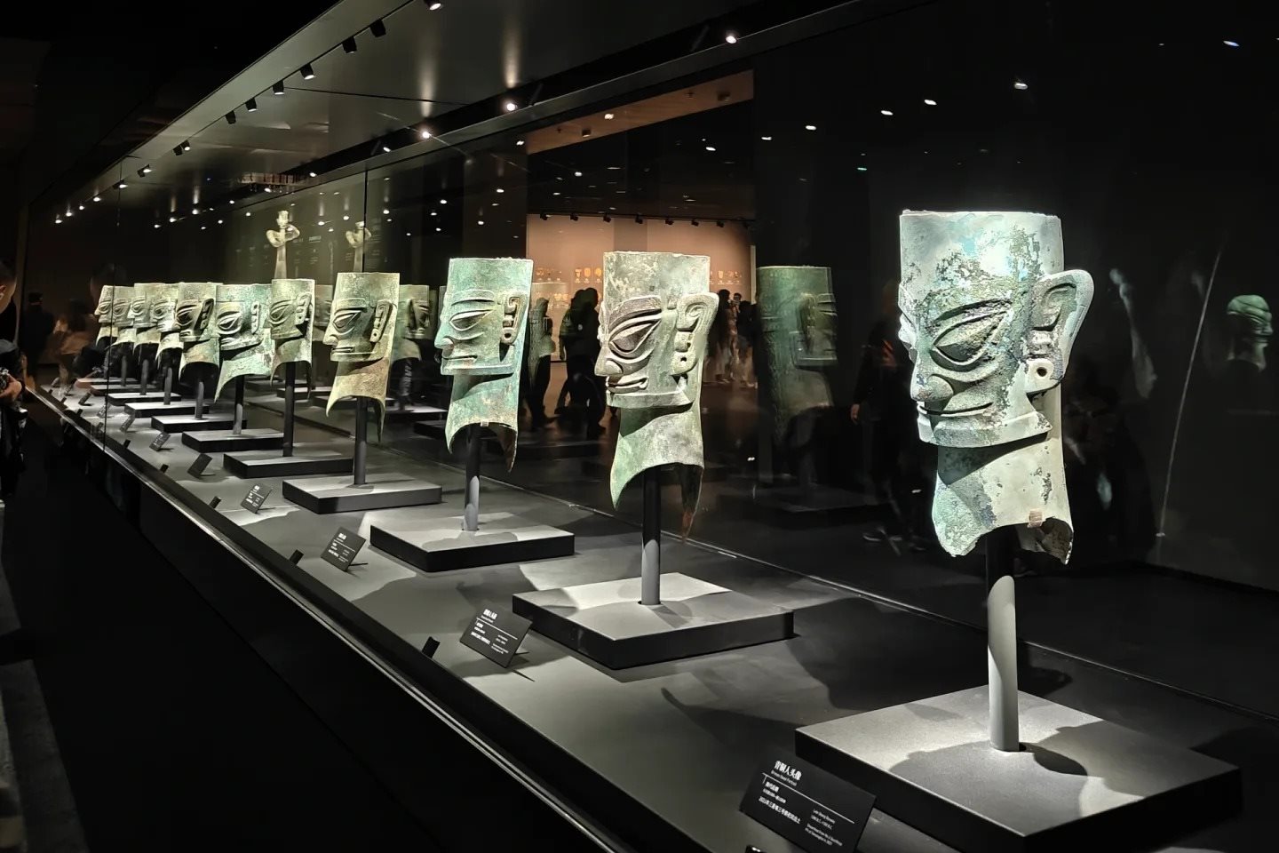 A long glass case running away from the camera, holding a row of more than a dozen bronze human heads on individual stands, each with angular features and wide eyes, lit from above against a dark hall.