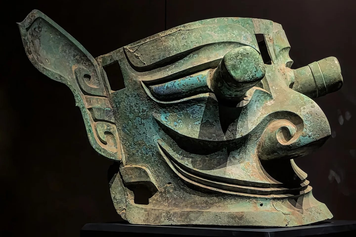 A large bronze mask lit against a black background in three-quarter view, showing cylindrical pupils projecting well forward of the eye sockets and wide flared ears, the metal green with patina.